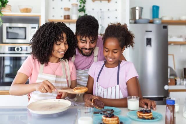 Glad positive young family foolishes with small daughter at kitchen, making pancakes, have tasty breakfast, pose at camera, drink fresh milk, eat pancakes