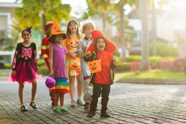 Child in Halloween costume. Mixed race kids and parents trick or treat on street. Little boy and girl with pumpkin lantern and candy bucket. Baby in witch hat. Autumn holiday fun.