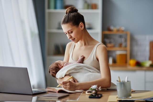 Portrait of focused Caucasian mother writing in notebook at table by laptop in living room while hugging her napping baby, copy space