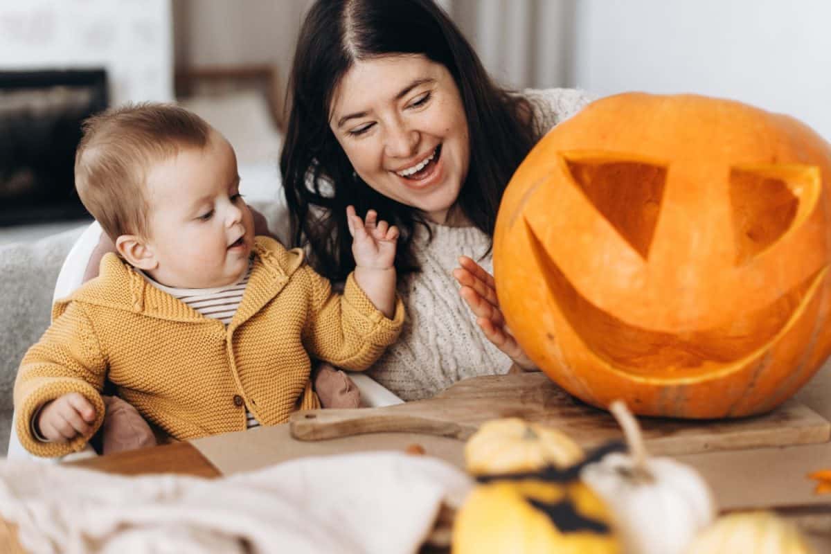Happy mother and cute baby son carving pumpkin on table with ghosts, spiders and bats decoration. Family cutting halloween pumpkin, making a jack o lantern. Halloween preparation