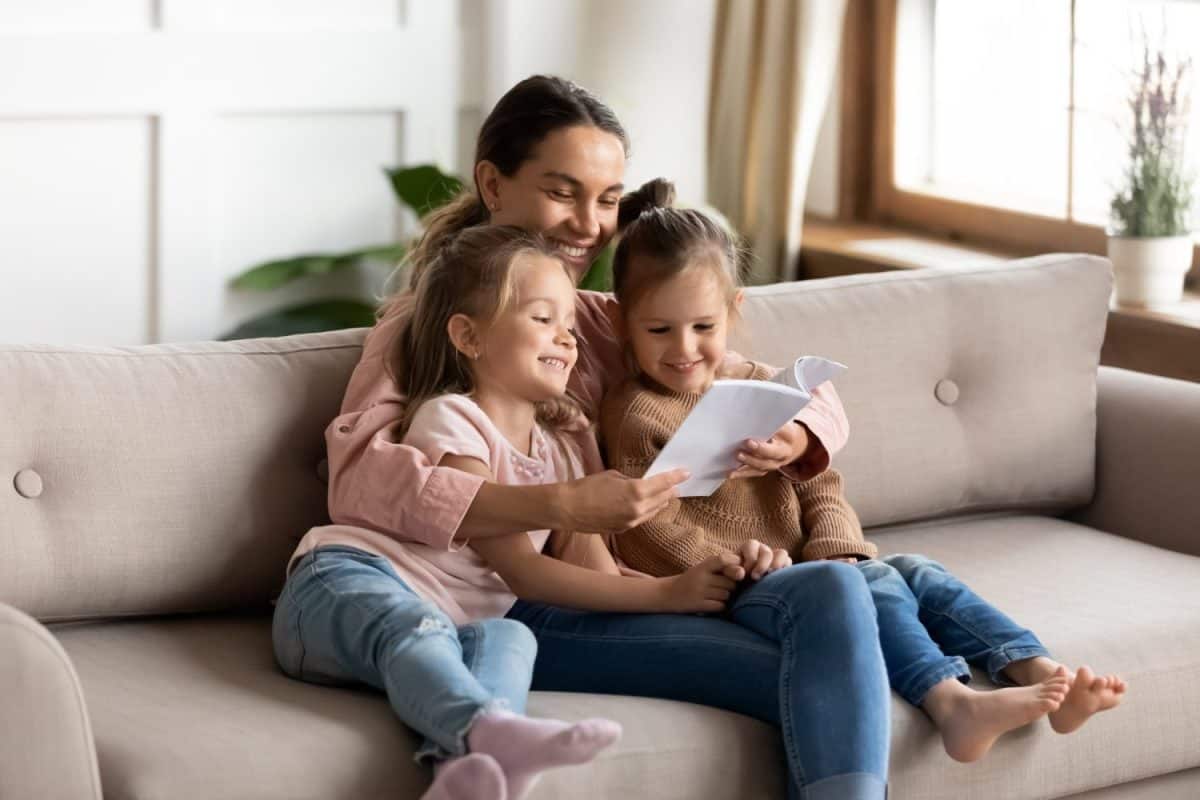 Happy family young mother babysitter hold read book relax embrace cute little children daughters, smiling parent mum tell small kids funny fairy tale story sit on sofa having fun together at home