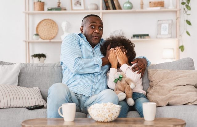 Scary movie night. Mature African American man with his little granddaughter closing face in horror at home