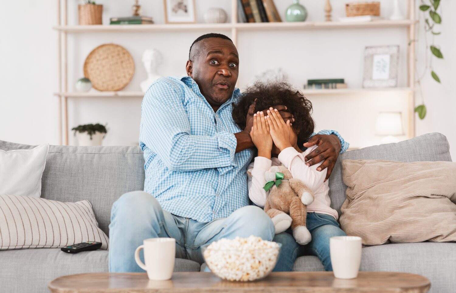 Scary movie night. Mature African American man with his little granddaughter closing face in horror at home