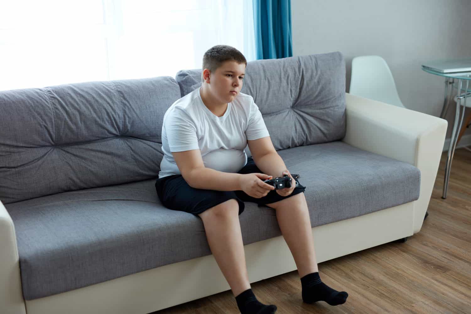 caucasian teenager boy enjoys video games, overweight boy sits on sofa holding console in hands, concentrated on game, look at screen