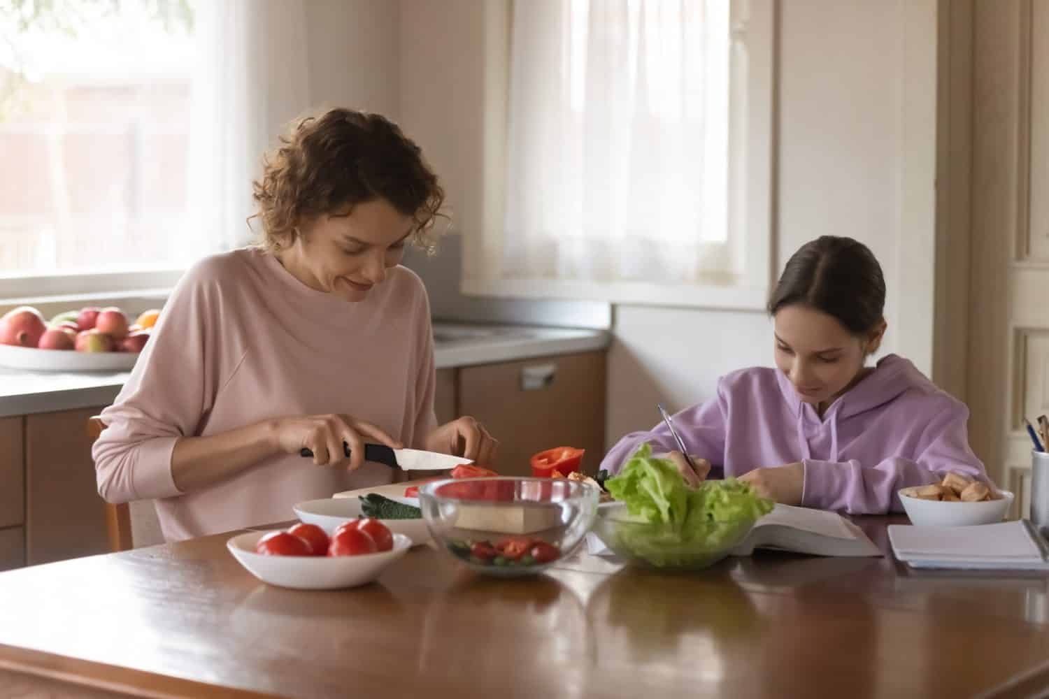 Happy Caucasian mother sit at table in kitchen cook prepare healthy food for family dinner. Smart teenage daughter schoolgirl study learn prepare homework assignment writing. Daily routine concept.