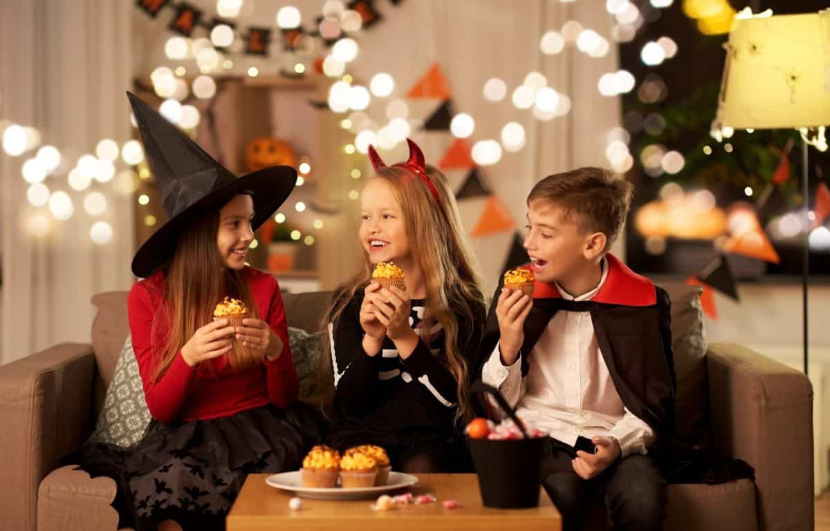 halloween, holiday and childhood concept - smiling boy and girls in costumes eating cupcakes and having fun at home at night