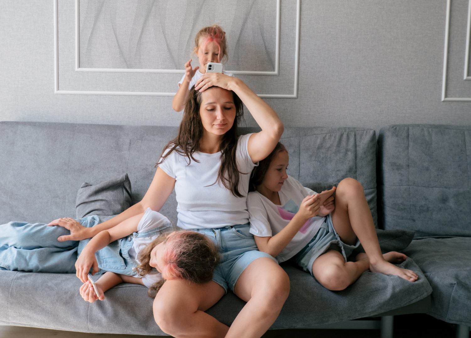 Tired young mother sits on couch and holds her head while her three naughty daughters play games on smartphones. lifestyle family together portrait.