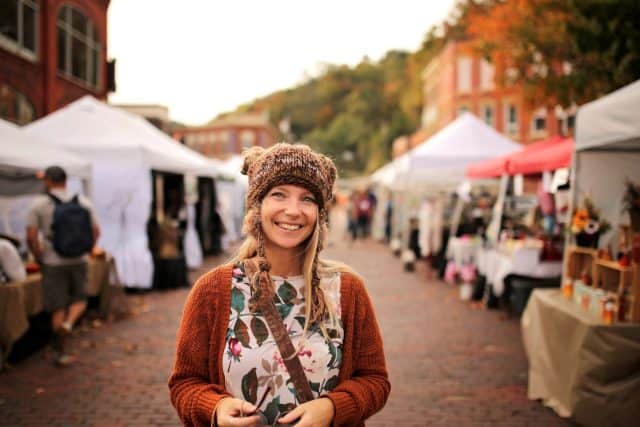 A happy women wearing a hand knit fox winter hat is smiling as she shops outside at a small town fall festival market.