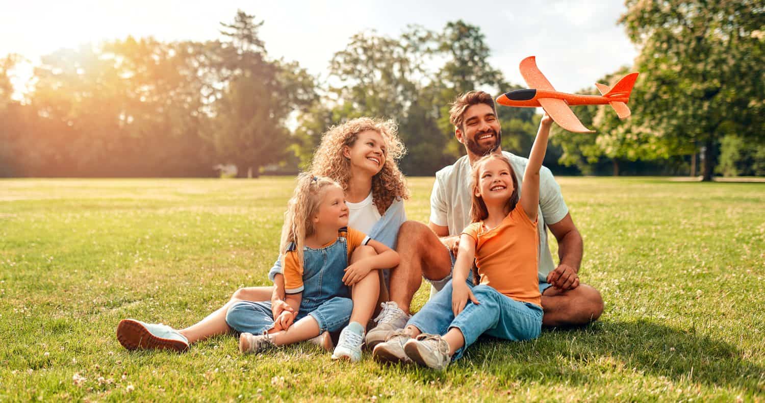 Happy young family dad, mom and two children daughters playing with an airplane sitting in a meadow on the grass in the park on a warm sunny day, having fun on a day off.