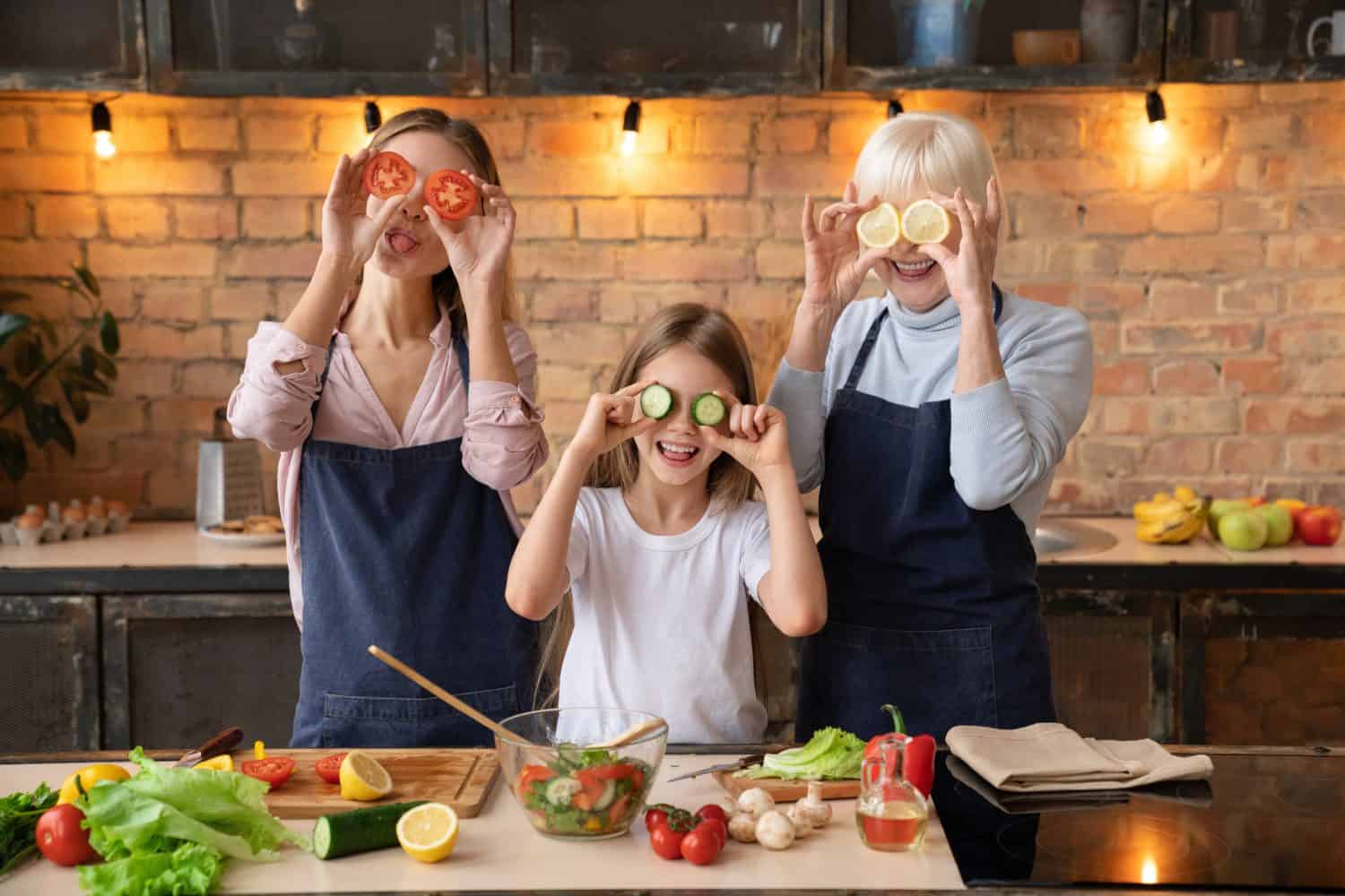 Young mother and her little cute daughter and grandmother having fun by putting pieces of organic fruit and vegetables to their eyes while cooking in kitchen. Happy family concept