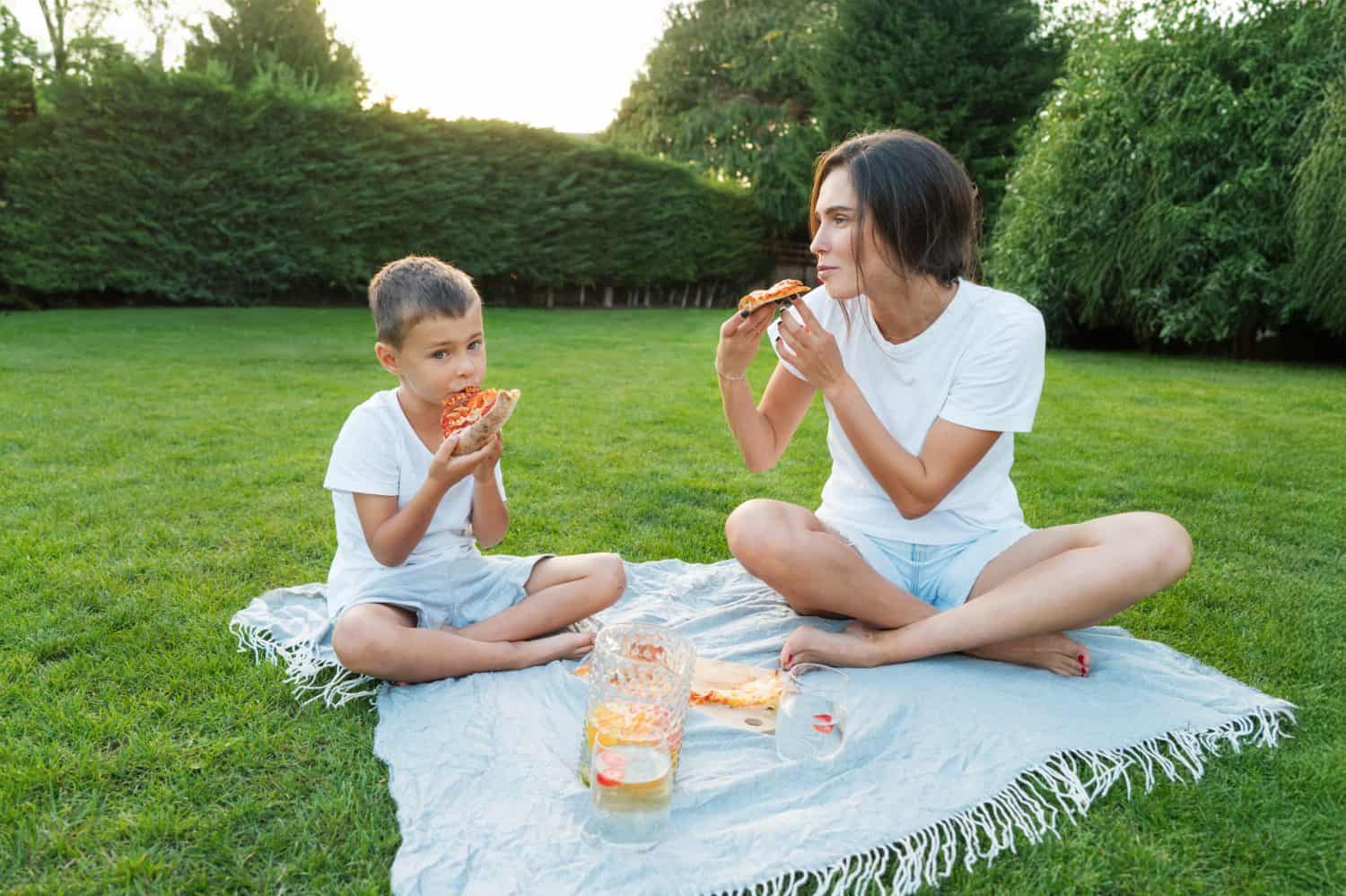Happy Young mother and preschooler son having outdoor picnic dinner, eating pizza sitting on backyard Lawn on Sunny Day. Happy family time together. Active childhood. House in the suburbs in summer.