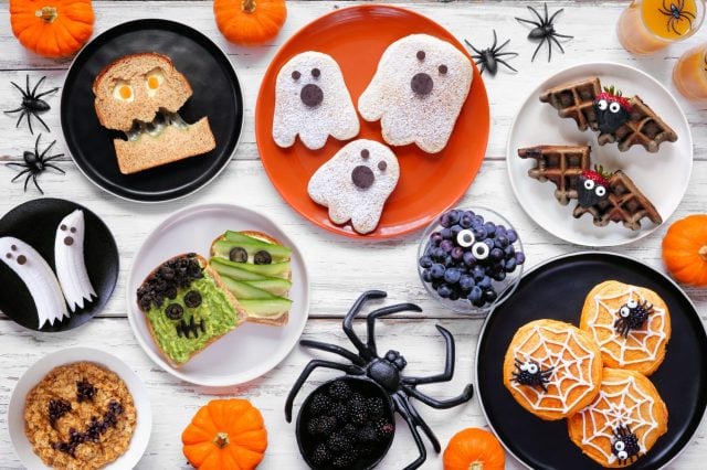 Spooky Halloween breakfast table scene over a white wood background. Top down view. Variety of pancakes, toast, oatmeal and fruit.