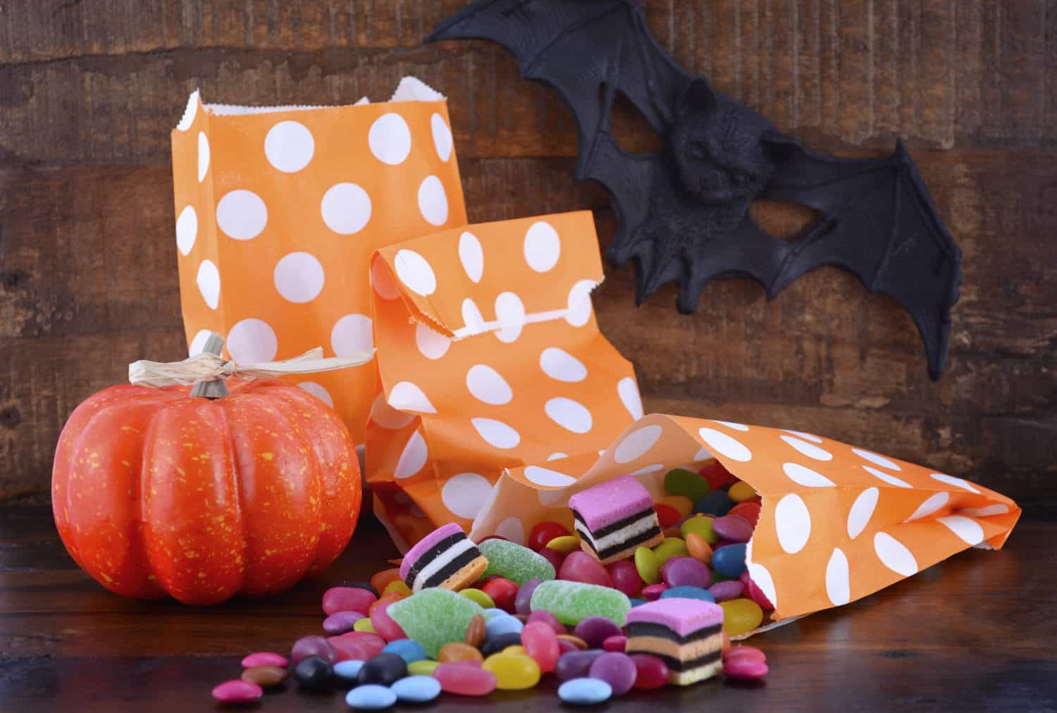 Halloween Candy Trick or Treat Bags with colorful candy, pumpkin and bat on dark wood table background.