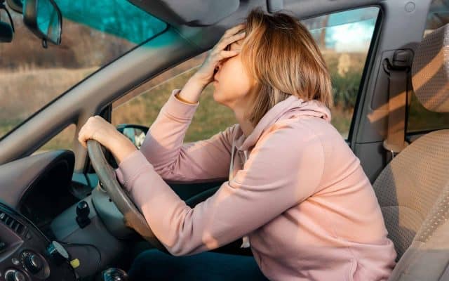 A young blonde sad woman is sitting behind the wheel of a car and holding her head in despair, covering her eyes with her hand. Side view. Stress while driving a right-hand drive car.