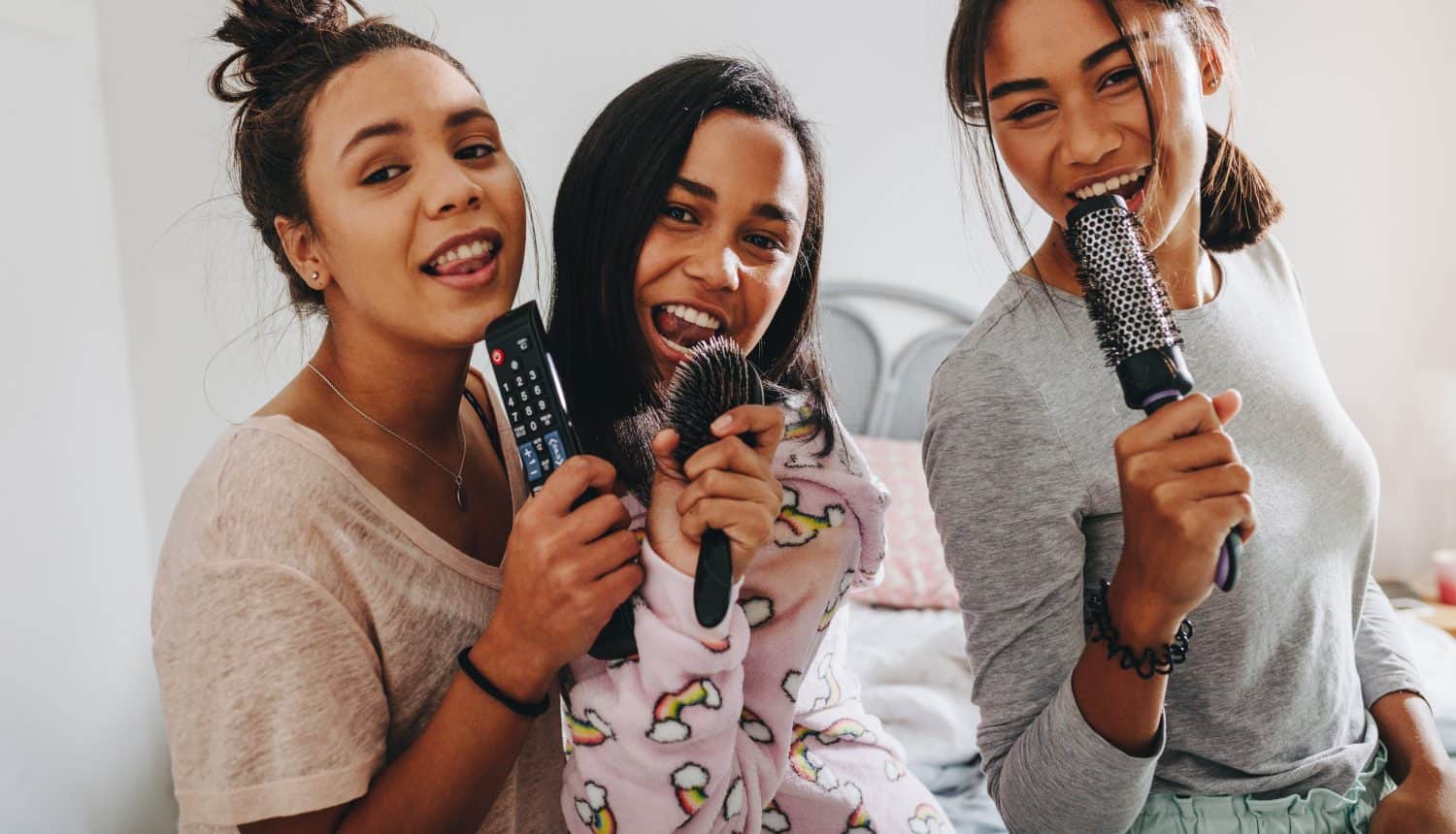 Happy young girls having fun singing songs in karaoke style holding hair brushes and remote control. Girls at a sleepover having fun singing together.