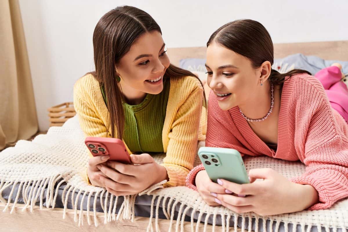 Two young women in casual attire lounging on a bed, engrossed in their cell phones.
