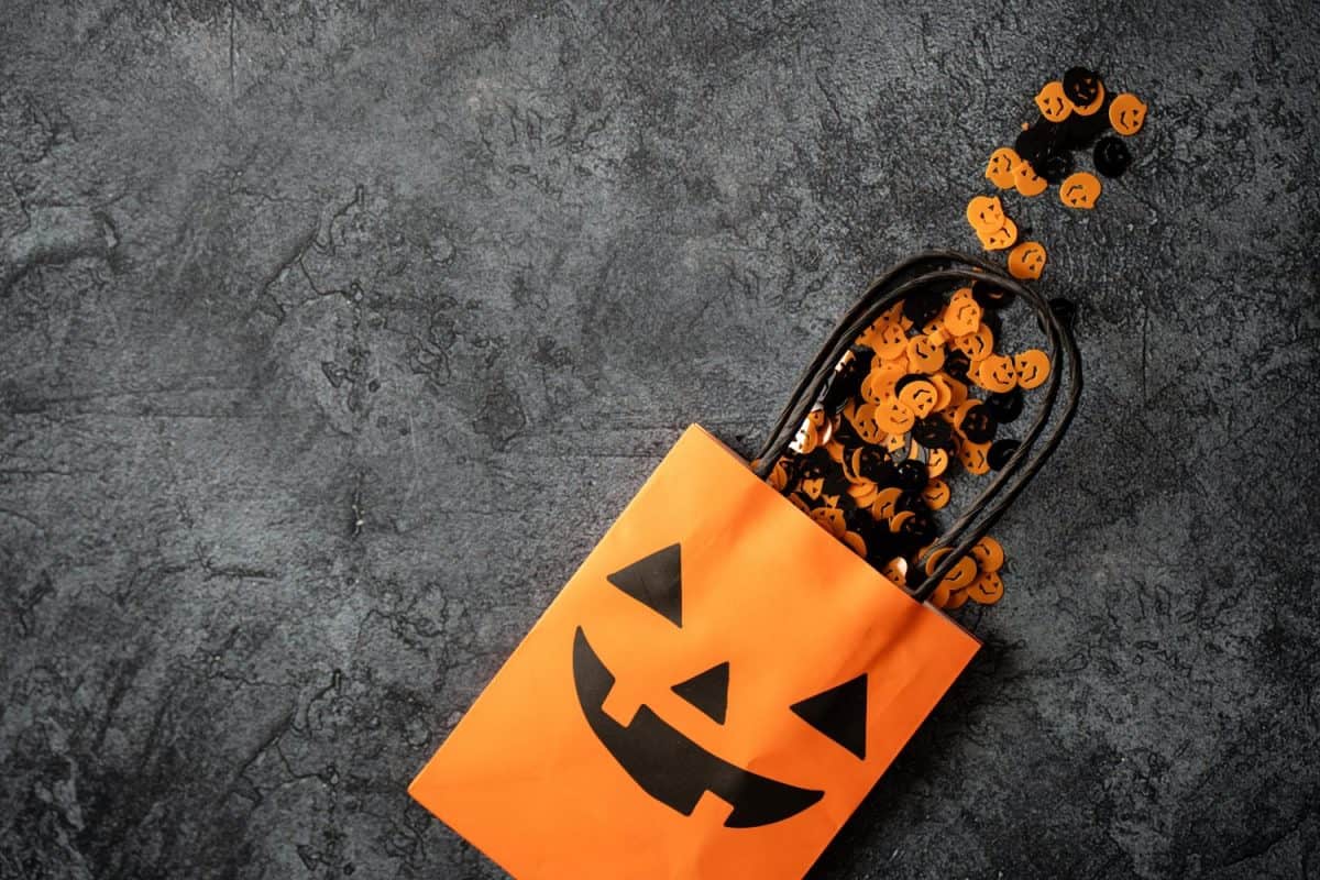 Pumpkin decorations spill from Halloween treat bag. Paper gift bag for trick-or-treating. Decorated with spooky Jack-o'-lantern face. Trick or Treat party favor bag. Composition with copy space.