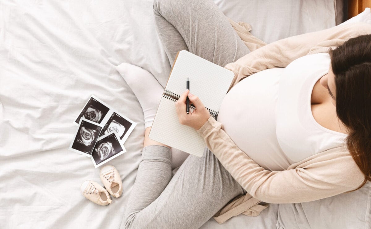 Pregnant woman making packing list for maternity hospital, preparing for labor at home, top view, empty space