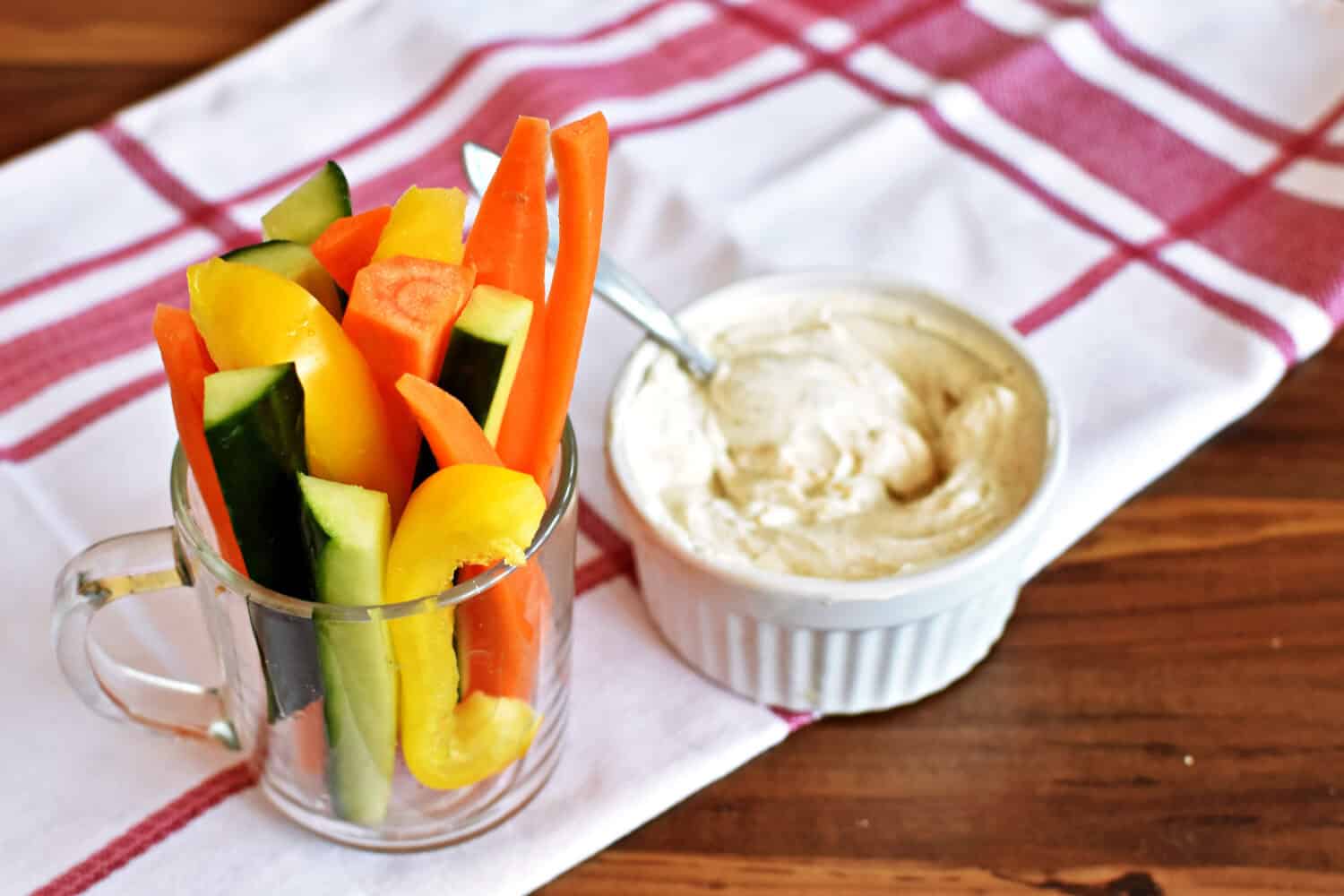 A jar full of carrot sticks, cucumber sticks and pieces of pepper stands on a dark wooden surface, next to it is a bowl of curd dip - delicious, healthy and fresh snack