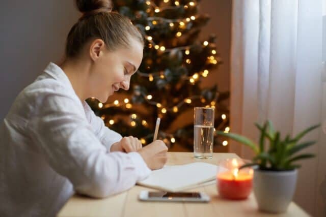 Indoor shot of young adult woman writing resolutions for New year in notebook at home, sitting by Christmas tree, wearing white shirt. Motivation for future.