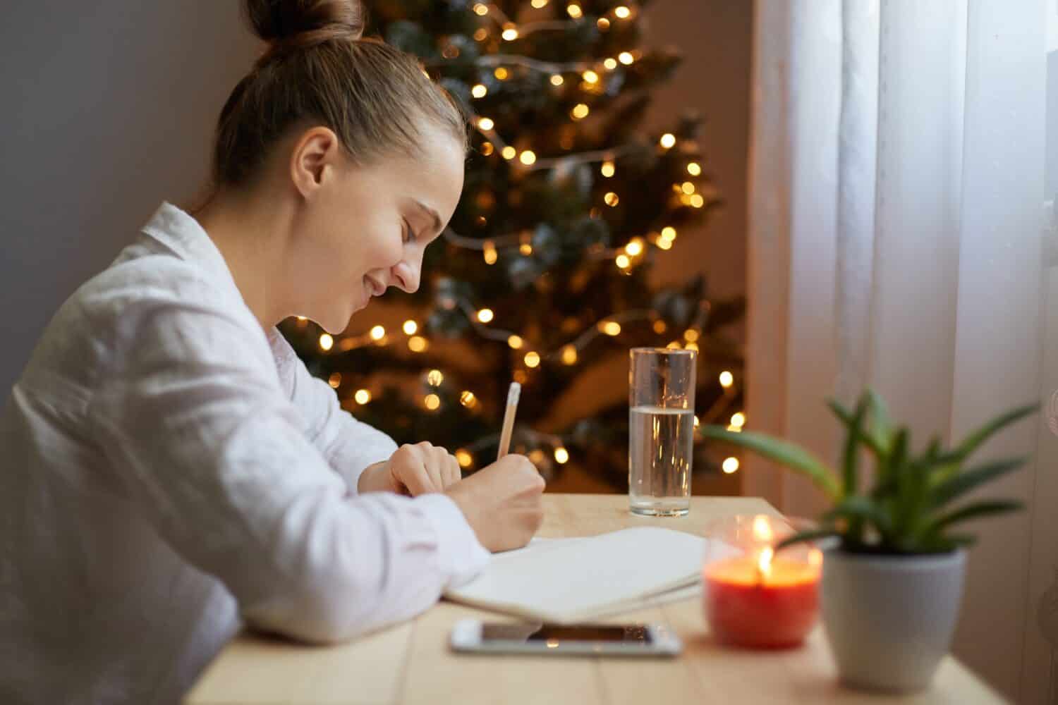 Indoor shot of young adult woman writing resolutions for New year in notebook at home, sitting by Christmas tree, wearing white shirt. Motivation for future.