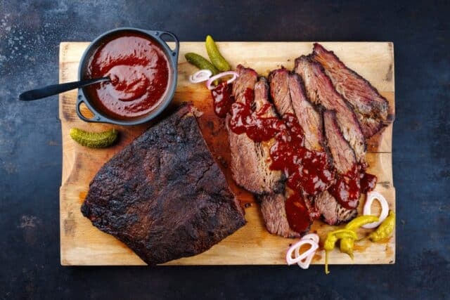 Traditional smoked wagyu beef brisket served with vegetable and spicy barbecue Louisiana sauce served as top view on a wooden cutting board