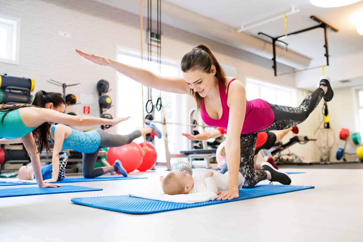 Mother smiling at baby while performing exercise above. Group exercise class, fitness or pilates for parents. Moms staying active while boding with babies.