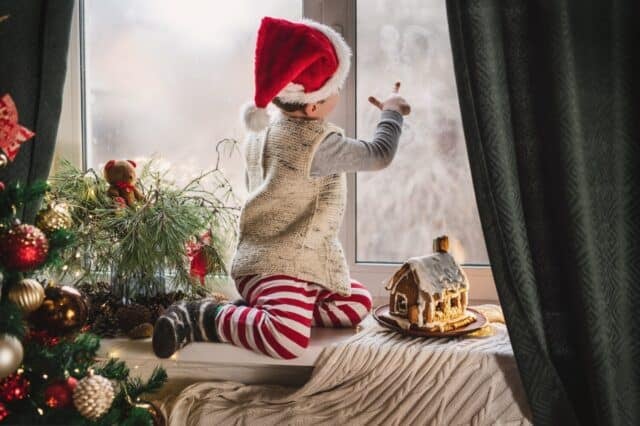 boy 3-4 years old in red and white stripped pajamas and Santa Claus hat plays on the window near the decorated New Year tree with lanterns and gingerbread house. Christmas symbols, holiday card