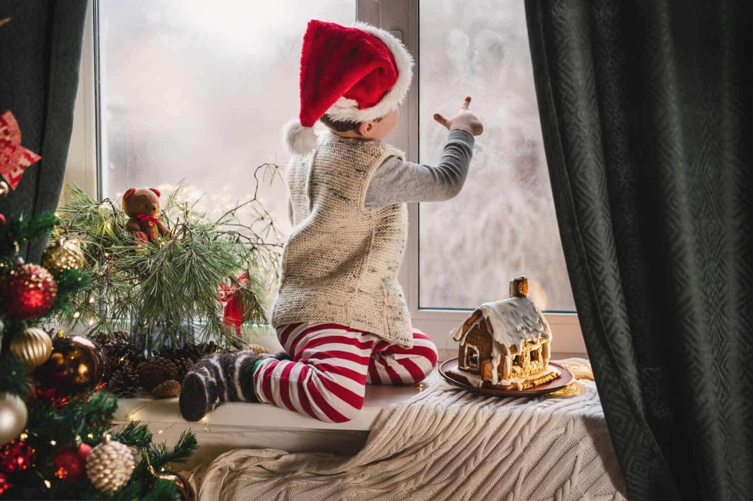 boy 3-4 years old in red and white stripped pajamas and Santa Claus hat plays on the window near the decorated New Year tree with lanterns and gingerbread house. Christmas symbols, holiday card