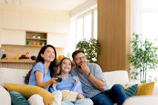 Three family members enjoy a joyful movie night, sitting on a comfortable couch filled with cushions, sharing laughter and popcorn during a cozy evening together.