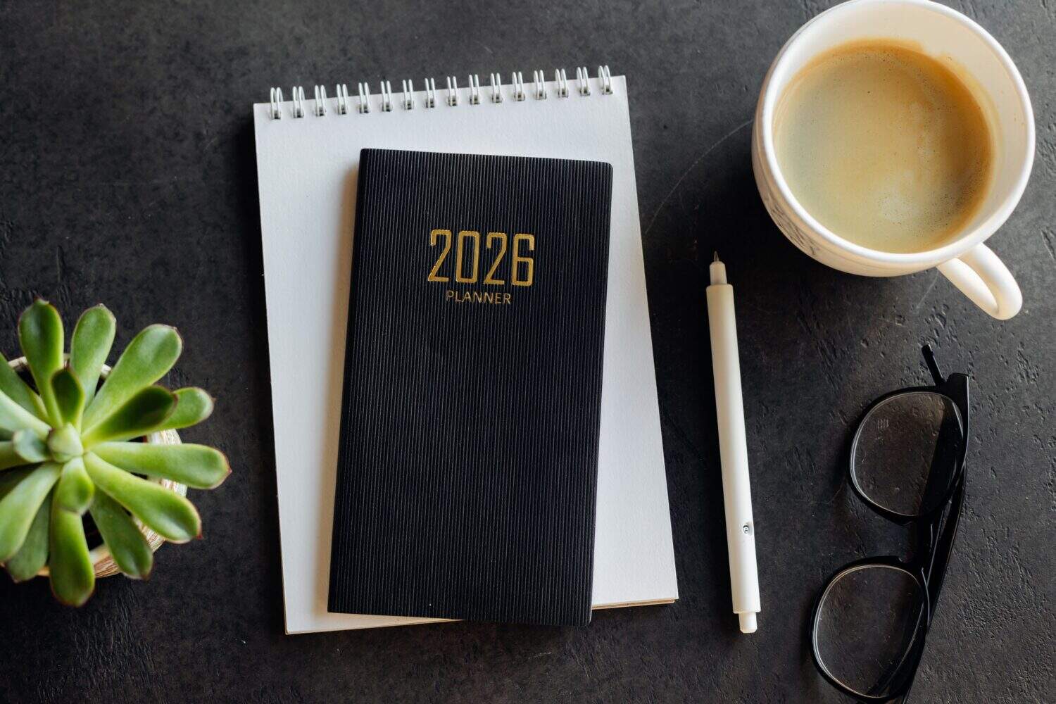 A tidy workspace featuring a 2026 planner, spiral notebook, white pencil, glasses, and a cup of coffee on a warm wooden desk, suggesting planning, focus, and productive mornings.