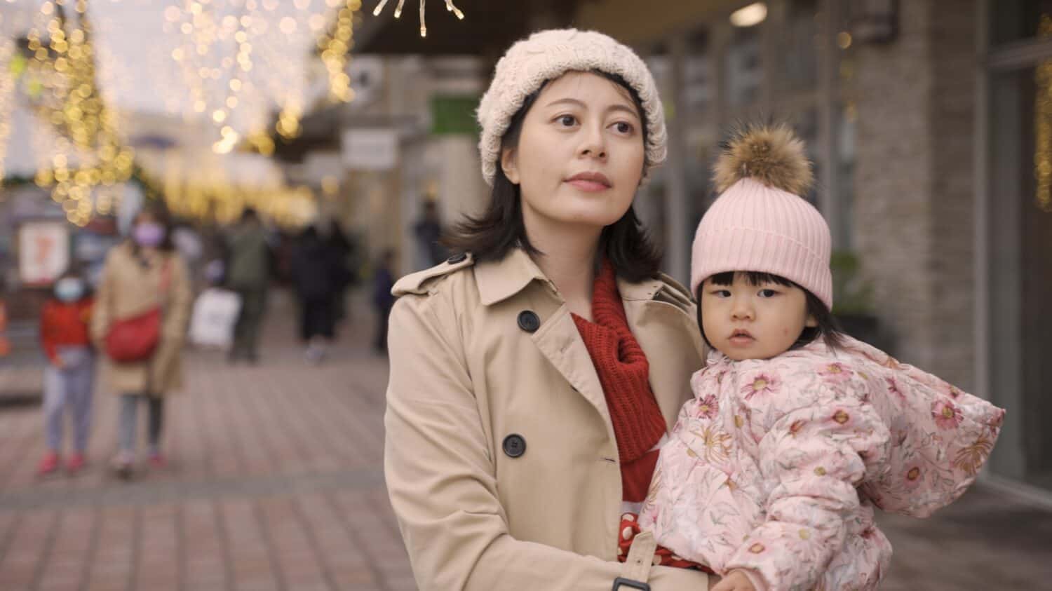 asian mother with her toddler baby in arms is looking at the beautiful cityscape on street with holiday lights at background during Christmas season