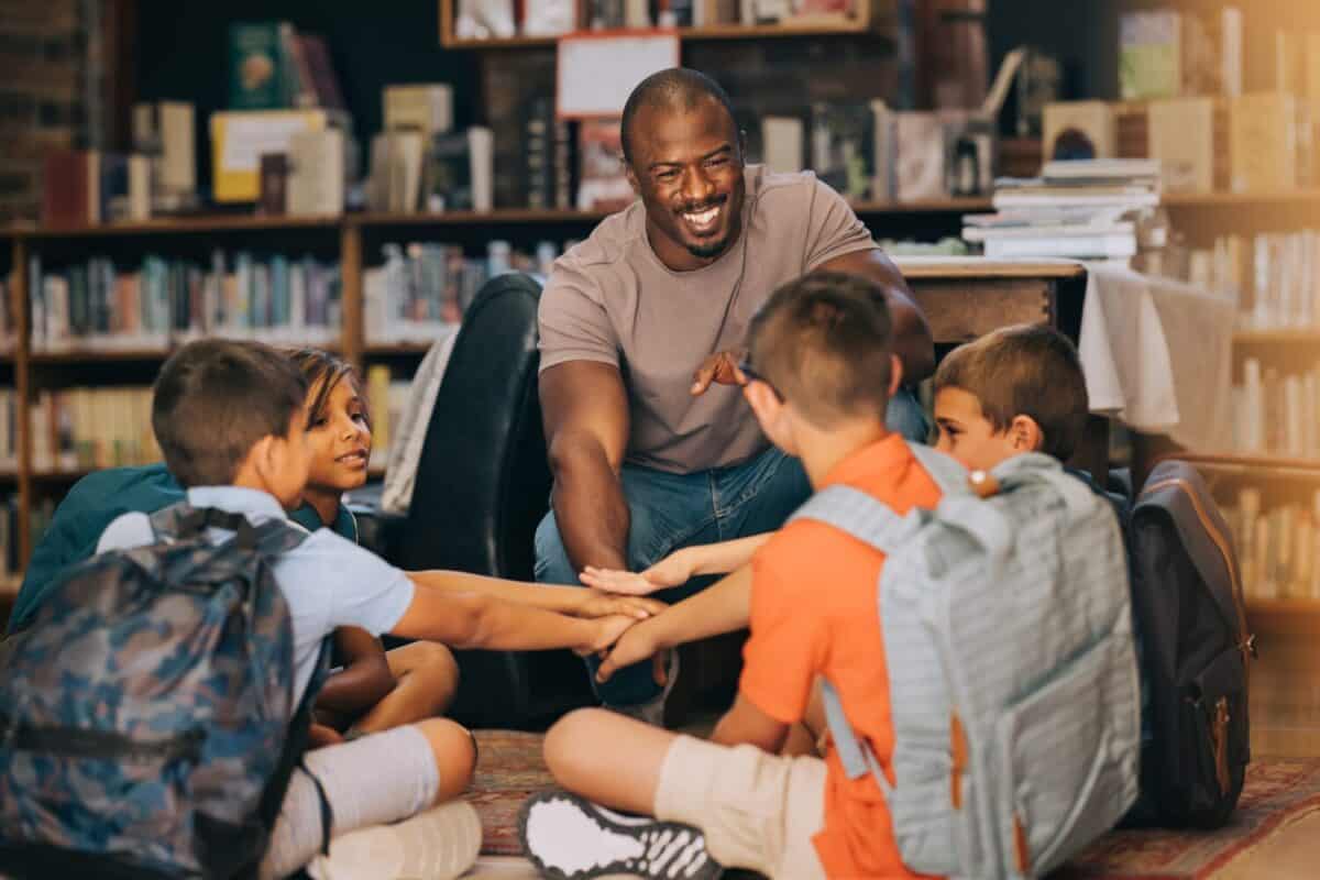 Male school teacher huddling with his students in a library. Man teaching a group of primary school children. Motivation and mentorship in elementary school.