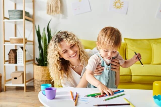 A curly-haired mother and her toddler daughter sit at a table, surrounded by crayons, as they engage in creative expression.