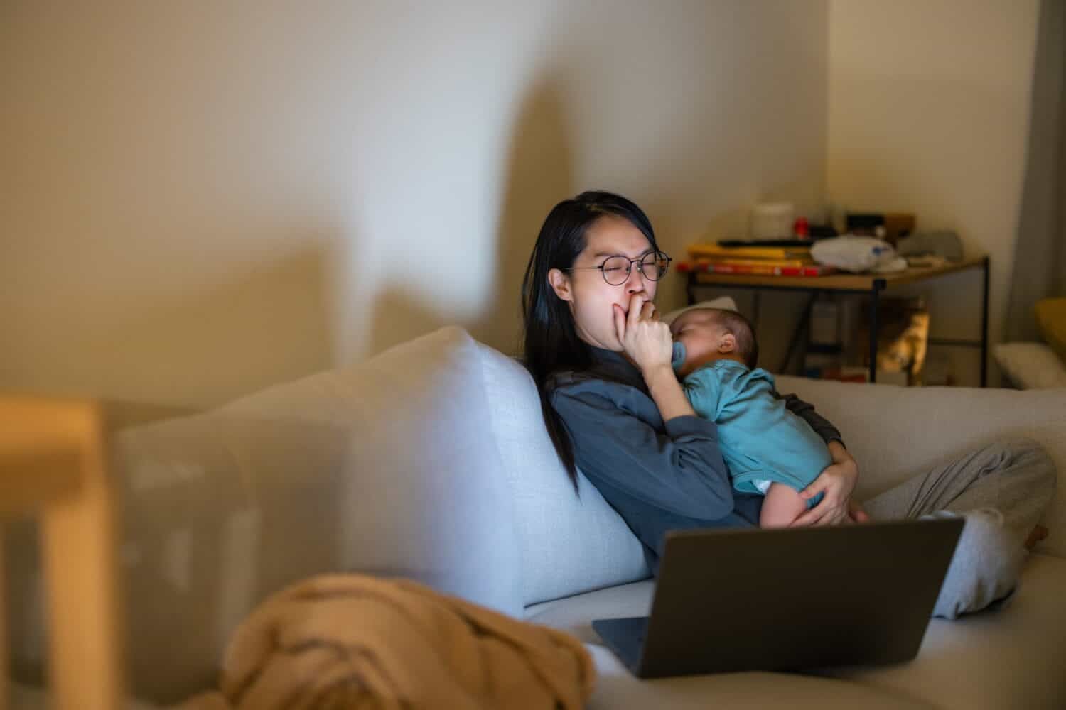 Tired working mother caring for baby while using laptop at home