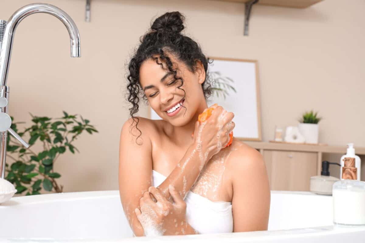 Beautiful young happy African-American woman taking bath with sponge and soap foam at home