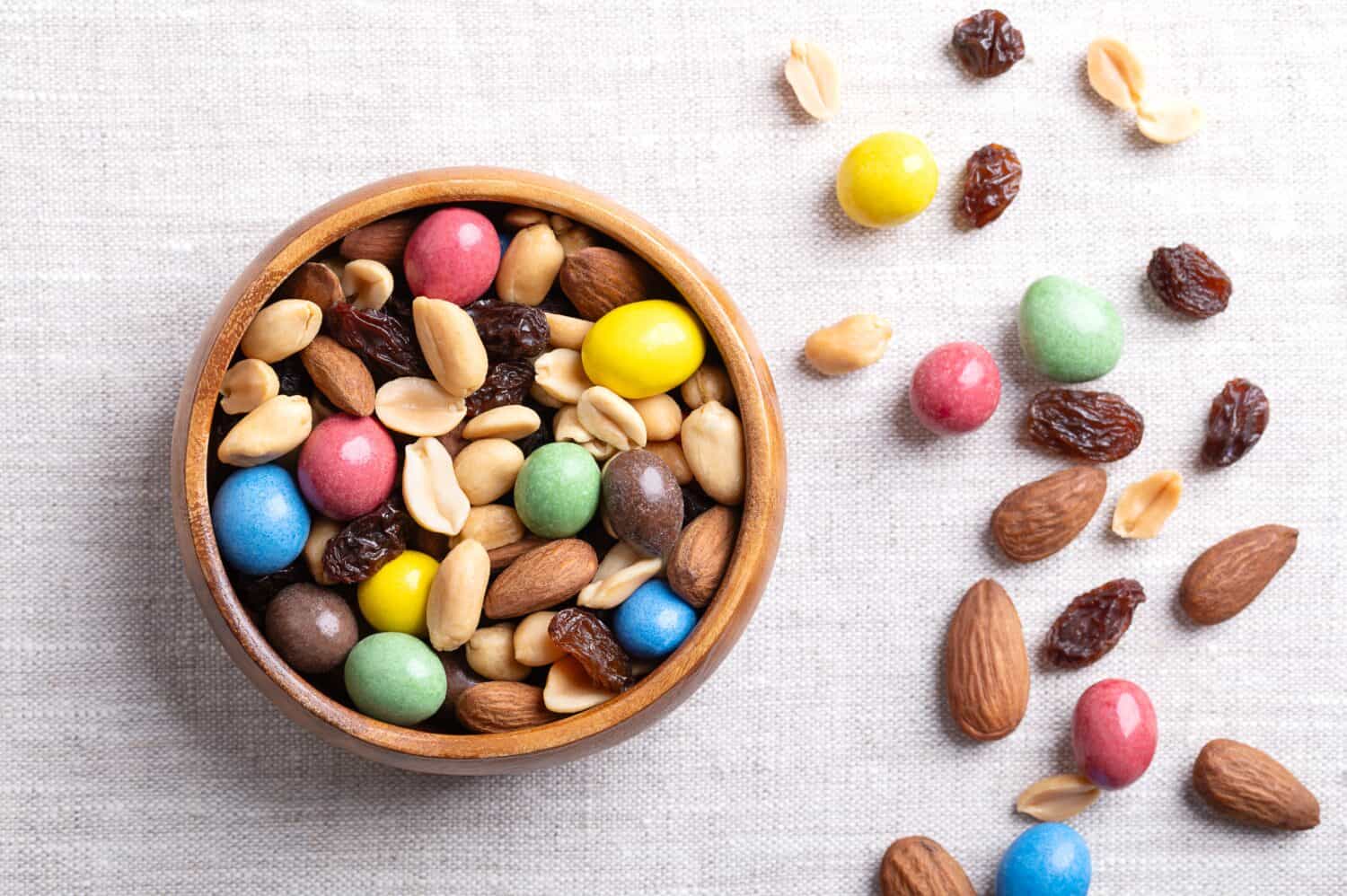 Rainbow trail mix in a wooden bowl on linen fabric. A snack mix of candy coated milk chocolate peanuts, roasted peanuts, raisins, and roasted almonds. A combination of sweets, dried fruits, and nuts.