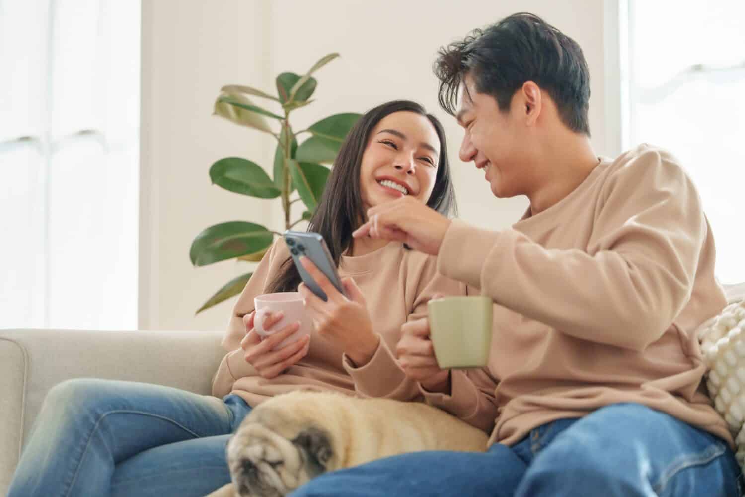 Smiling Asian man and woman relaxing on sofa with their dog, looking at smartphone and sharing joy and affection in home.