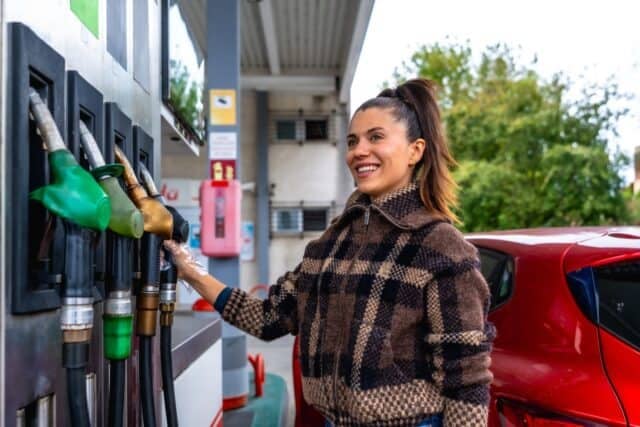 Woman smiling and wearing a protective glove while selecting a fuel pump to refuel her red car at a gas station, showcasing daily tasks and vehicle maintenance