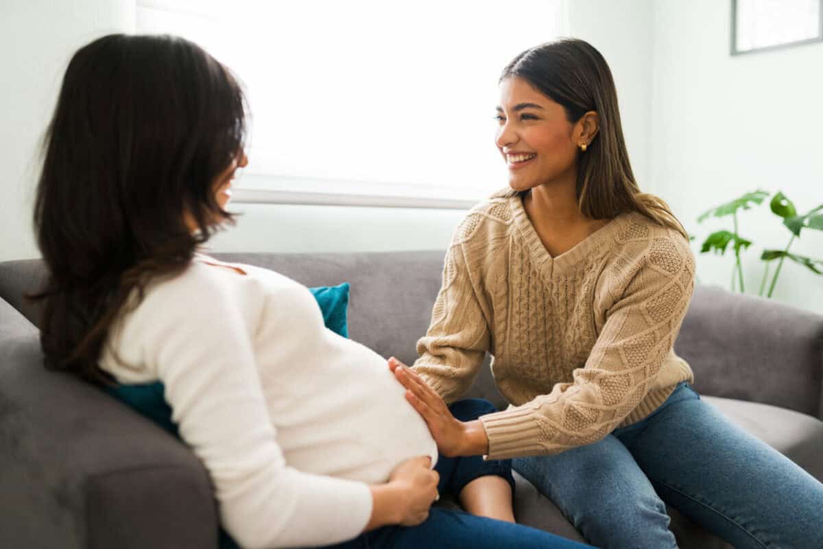 Expectant mother sitting on the sofa during a home visit from her happy doula. Smiling midwife touching the belly of a pregnant woman