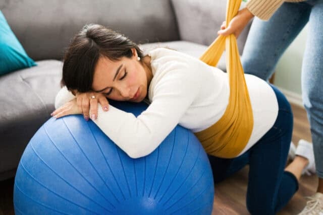 Caucasian pregnant woman doing breathing exercises and resting on a fitness ball while her doula wraps a rebozo around her round belly