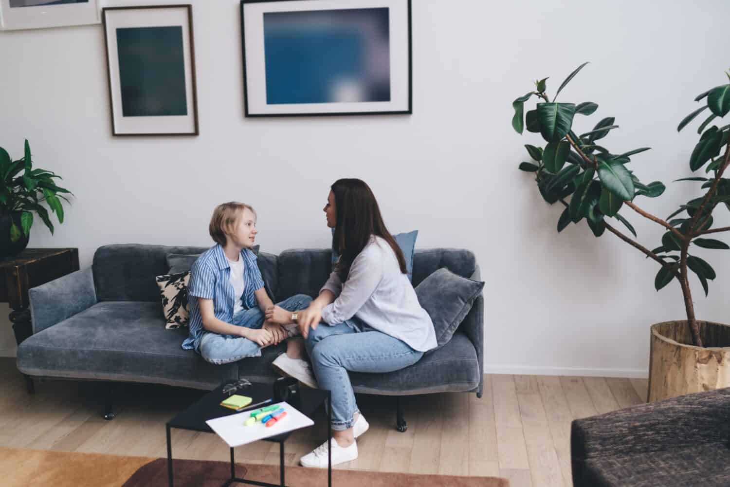 Calm mother and preteen son in casual clothes sitting on comfortable sofa together and looking at each other while having conversation