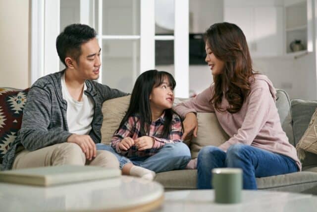 young asian mother and father sitting on family couch at home having a pleasant conversation with daughter