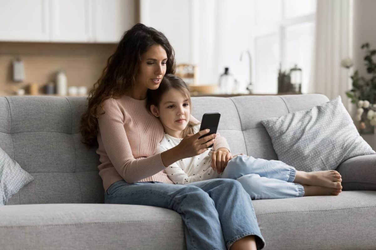 Positive beautiful brunette mom teaching sweet child girl to use online service on smartphone, relaxing on comfortable home sofa, holding mobile phone, hugging kid