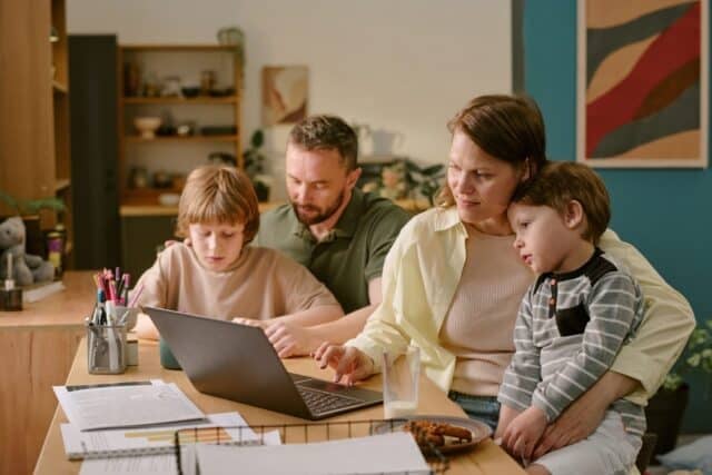 Caucasian middle aged woman working on laptop with young Caucasian boy on lap, Caucasian middle aged man assisting Caucasian child with homework, family multitasking during work from home