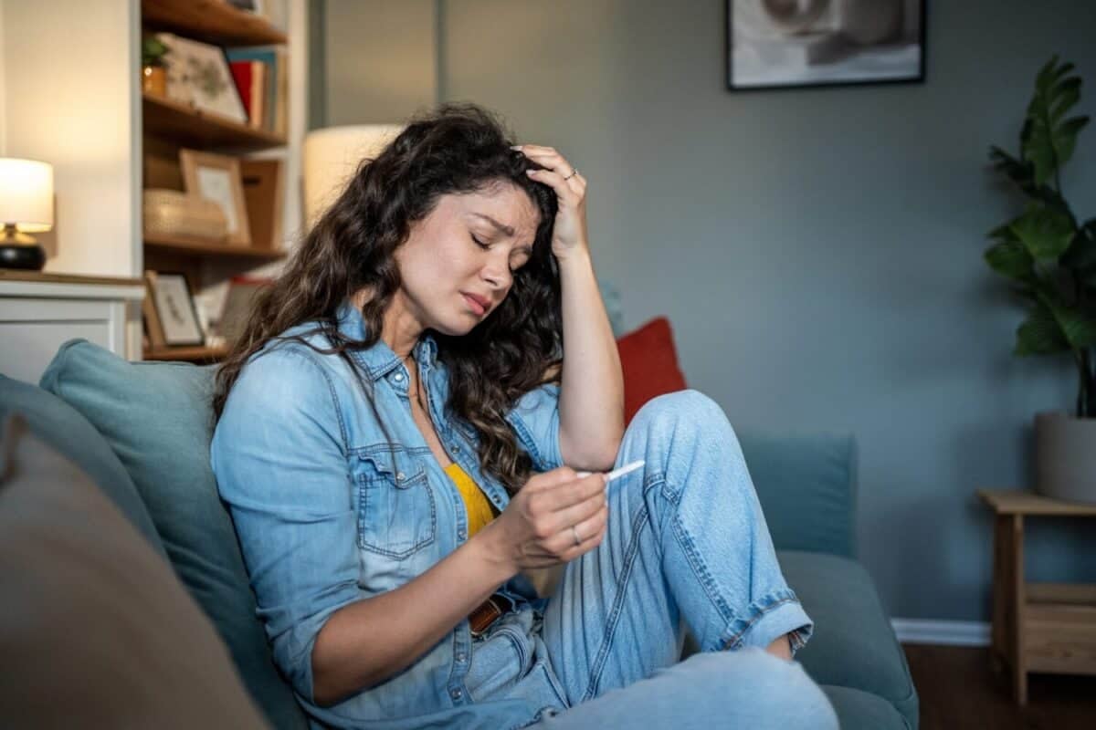 Sad woman is holding positive pregnancy test and touching her forehead while sitting on sofa at home, suffering from unwanted pregnancy and feeling desperate and worried about future