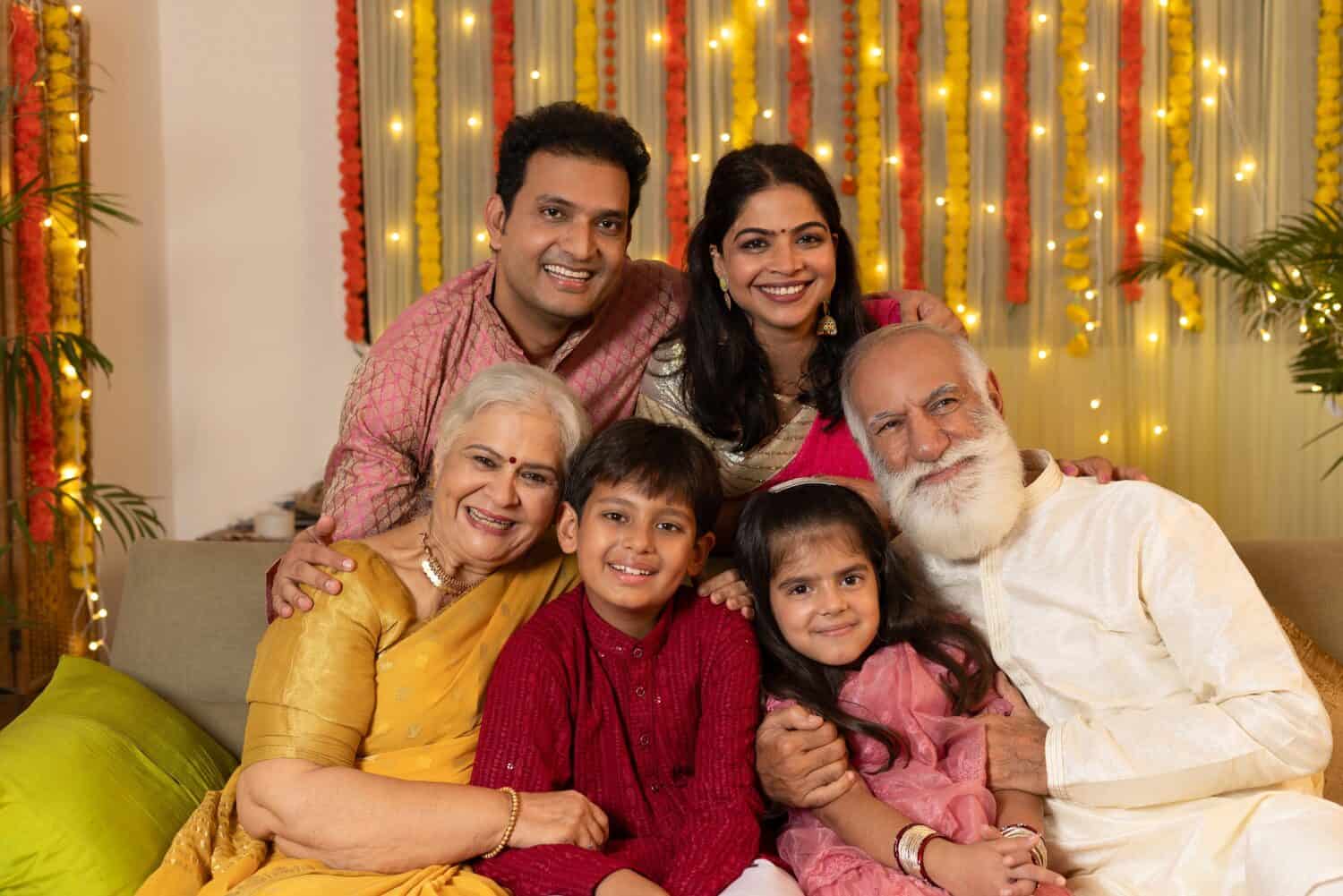 Indian happy family celebrating Diwali festival in festive season while siting together and looking at the camera at home - colorful decorated background - Diwali holidays festive