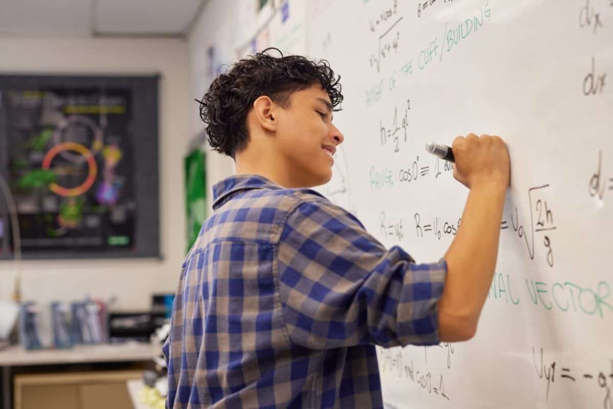 Happy multiethnic male student writing math equation on the whiteboard at high school. Smiling focused teenager solving physics formulas on a white board with erasable marker.