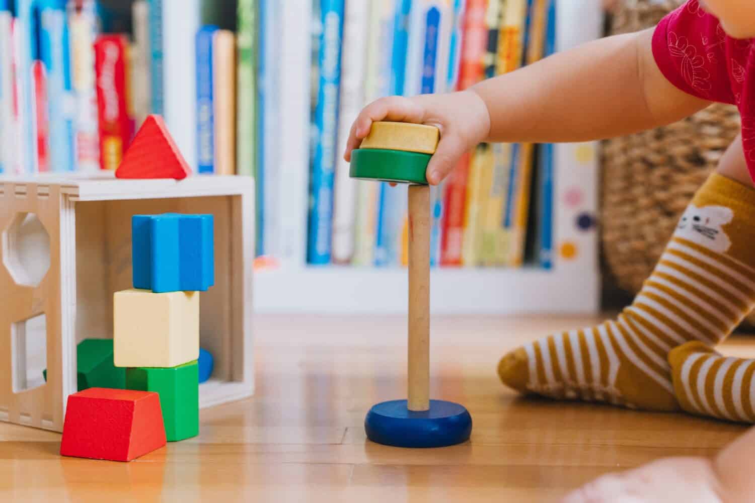 Toddler playing with wooden toys indoors. Baby sitting on floor throwing toys around and exploring shapes and colors. Montessori education. Motor skills, early preschool learning concepts.