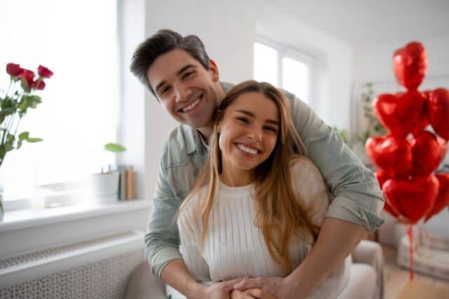 Happy young couple hugging and smiling at home, celebrating Valentine’s Day together. Romantic relationship, love, lifestyle and cozy modern interior with heart shaped balloons.